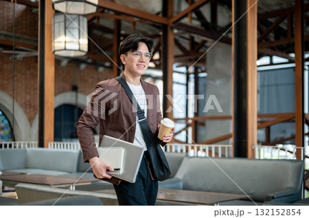 Brown suit man with glasses walking while holding a coffee cup with laptop and notebook in the cafe. Brown suit man with glasses walking while holding a coffee cup with laptop and notebook in the cafe. 132152854