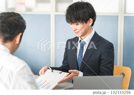 資料を見せながらショールーム・商談スペースで接客する若いアジア人ビジネスマン 資料を見せながらショールーム・商談スペースで接客する若いアジア人ビジネスマン 132153256