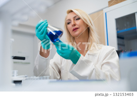 Middle aged female scientist watching chemical solution in glass flask in laboratory 132153403