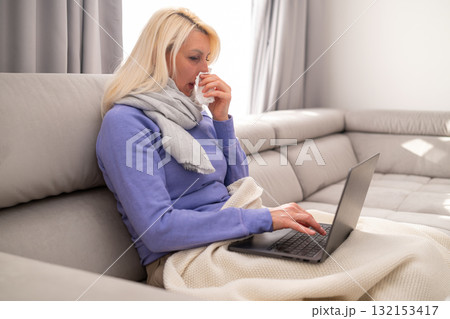 Middle aged blonde woman sitting on couch with a blanket, feeling unwell and watching movies on her laptop at home. Concept of illness, rest, relaxation, and cozy home atmosphere. 132153417