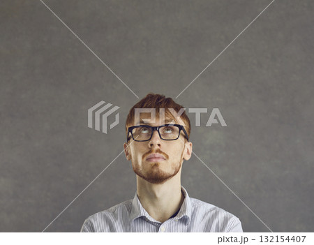 Young man with a questionable expression looks up while standing on a gray concrete background. Young man with a questionable expression looks up while standing on a gray concrete background. 132154407