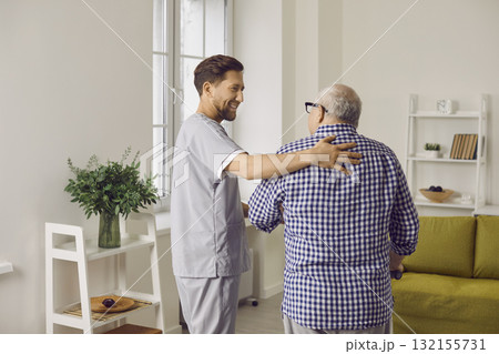 Smiling young male caregiver in uniform hugging elderly man during visit Smiling young male caregiver in uniform hugging elderly man during visit 132155731