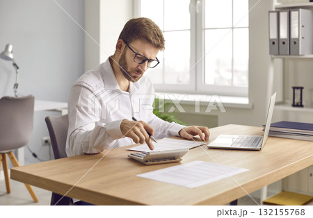 Young focused serious business man working on laptop at office making calculations. 132155768