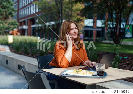 Plus size woman working remotely from coffee shop, making phone call with client. 132156047