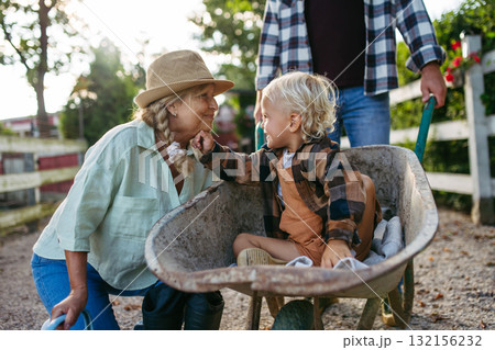 Grandmother playing with grandson in a wheelbarrow on a family farm. 132156232