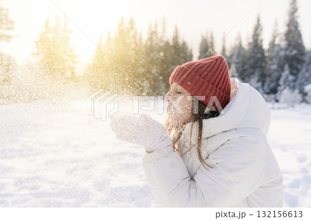 A young woman dressed warmly in a white jacket and red knitted hat stands in a snowy forest. She joyfully blows snowflakes into the air, creating sparkling particles illuminated by the sunlight. A young woman dressed warmly in a white jacket and red knitted hat stands in a snowy forest. She joyfully blows snowflakes into the air, creating sparkling particles illuminated by the sunlight. 132156613