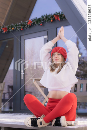A woman in white clothes and a red cap sits in the lotus position in winter and practices yoga. Perhaps she is on winter vacation outside the city during the winter holidays and does sports. 132156628