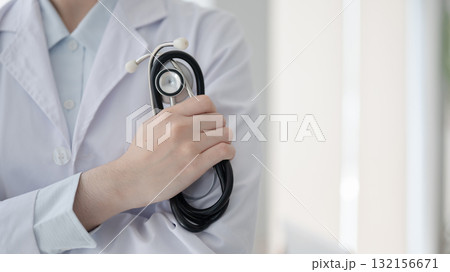 Close up of a doctor hand holding a stethoscope while standing with arms crossed in diagnosis room. Close up of a doctor hand holding a stethoscope while standing with arms crossed in diagnosis room. 132156671
