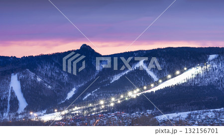 Evening panorama mountain ski resort. illuminated ski slope with burning lights during a beautiful lilac sunset. A serene and tranquil twilight settles over the picturesque snowy mountains, showcasing Evening panorama mountain ski resort. illuminated ski slope with burning lights during a beautiful lilac sunset. A serene and tranquil twilight settles over the picturesque snowy mountains, showcasing 132156701