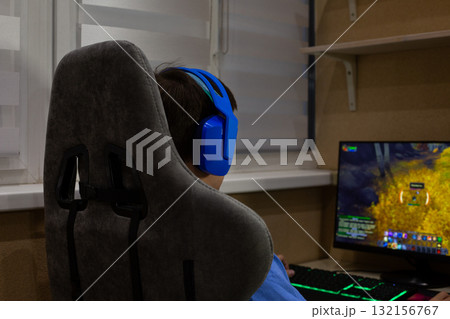 Young gamer wearing blue headphones sits at desk, playing an online video game on computer. The image captures the immersive experience of modern gaming, with vivid on screen action 132156767