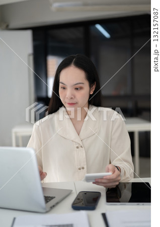Female office worker is holding a calculator and looking at laptop while sitting at table in office. Female office worker is holding a calculator and looking at laptop while sitting at table in office. 132157087