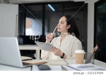 Businesswoman or office worker thinking while holding stylus and tablet sitting at table in office. 132157094