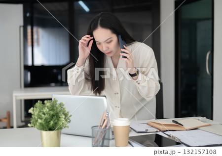 Female office worker sweeping her hair and talking on phone while leaning to look at laptop on table Female office worker sweeping her hair and talking on phone while leaning to look at laptop on table 132157108