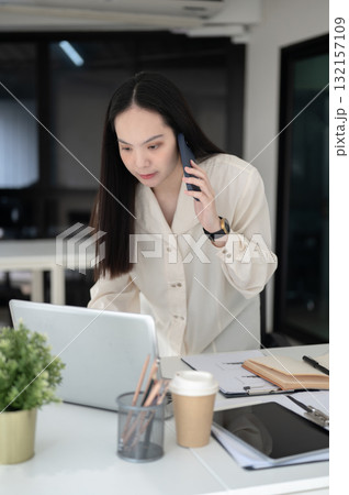 Businesswoman or female office worker is talking on phone while leaning to look at laptop on table. Businesswoman or female office worker is talking on phone while leaning to look at laptop on table. 132157109