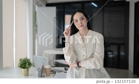 Female office worker holding tablet with stylus touching her head standing in front of working table 132157110