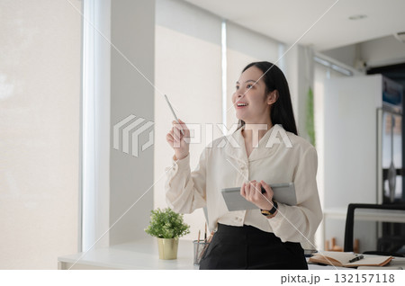 Female office worker smiling and looking away from tablet while standing in front of working table. Female office worker smiling and looking away from tablet while standing in front of working table. 132157118