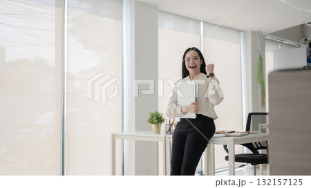 Female office worker smiling happily and raising a fist while hugging laptop and leaning on table. Female office worker smiling happily and raising a fist while hugging laptop and leaning on table. 132157125