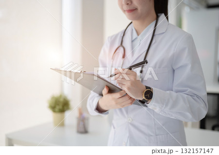 Female doctor with stethoscope writing with a pen on clipboard standing near table in diagnosis room 132157147