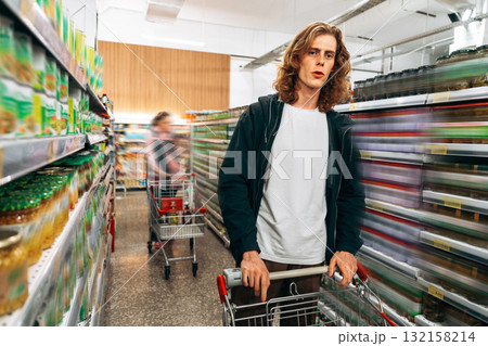 Young man shopping in a grocery store aisle with a cart and blurred background activity Young man shopping in a grocery store aisle with a cart and blurred background activity 132158214