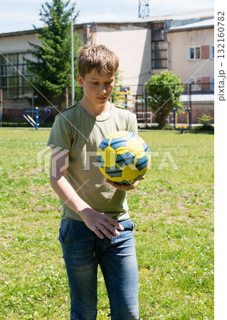 Teenager holding a soccer ball outdoors on a sunny day. He's near a building and some trees. 132160782
