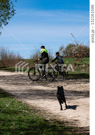 Two cyclists enjoying a sunny day ride on a dirt road, with a dog following. Two cyclists enjoying a sunny day ride on a dirt road, with a dog following. 132161086