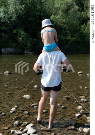 A child enjoys a piggyback ride through a shallow river, the water glistening around smooth stones. 132161093