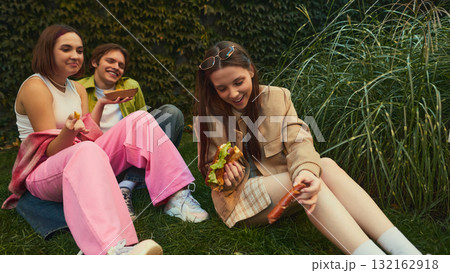 Young friends relaxing on grass laughing and eating burgers together Young friends relaxing on grass laughing and eating burgers together 132162918