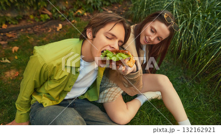 Boy taking big bite of burger while girl smiles watching him eat Boy taking big bite of burger while girl smiles watching him eat 132162921