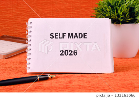 On a vibrant orange surface, a notebook titled Self Made rests beside a decorative plant and a sleek pen. This setup encourages creativity and inspires ambitious plans for the year 2026 132163066