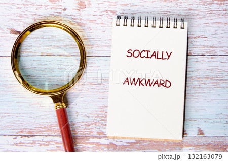 A notepad displays the words socially awkward, accompanied by a golden magnifying glass, all resting on a rustic wooden surface that adds charm to the scene, encouraging introspection A notepad displays the words socially awkward, accompanied by a golden magnifying glass, all resting on a rustic wooden surface that adds charm to the scene, encouraging introspection 132163079