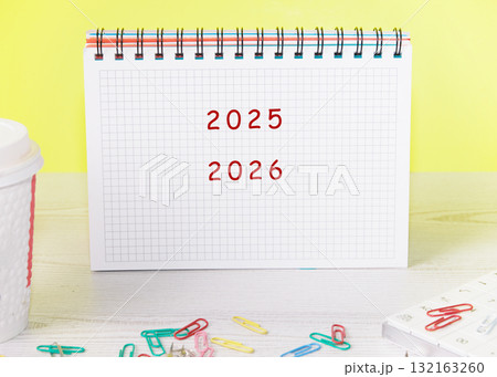 A calendar is opened to showcase the years 2025 and 2026. Colorful paper clips scattered around add a playful touch to this organized workspace against a cheerful backdrop A calendar is opened to showcase the years 2025 and 2026. Colorful paper clips scattered around add a playful touch to this organized workspace against a cheerful backdrop 132163260