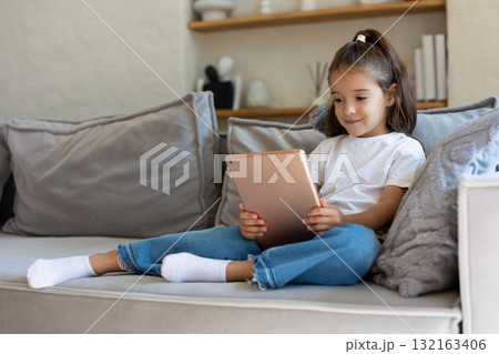 Preschool girl enjoying app on tablet while sitting on sofa Preschool girl enjoying app on tablet while sitting on sofa 132163406