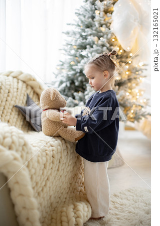 A young girl in a cozy setting interacts with a teddy bear near a decorated Christmas tree, creating a warm and festive atmosphere. A young girl in a cozy setting interacts with a teddy bear near a decorated Christmas tree, creating a warm and festive atmosphere. 132165021