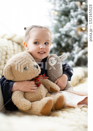 A young girl smiles while holding a teddy bear and a llama plush toy, with a Christmas tree in the background. 132165028