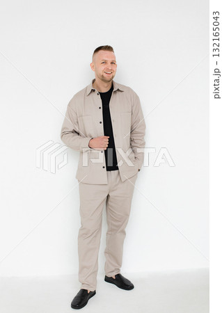 A smiling man stands in a studio setting, wearing a beige shirt and pants, paired with a black t-shirt and shoes. 132165043