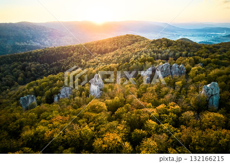 Aerial view of sun sets behind lush, green forest with dramatic rock formations. Warm light bathes landscape in golden hue. Dovbush Rocks, Carpathian mountains, Ukraine. 132166215