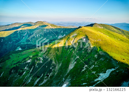 Aerial view of rugged mountain range with lush green slopes at sunset. Landscape showcases beauty of untouched nature with distant peaks and valleys bathed in soft sunlight. Carpathians, Chornogora. Aerial view of rugged mountain range with lush green slopes at sunset. Landscape showcases beauty of untouched nature with distant peaks and valleys bathed in soft sunlight. Carpathians, Chornogora. 132166225