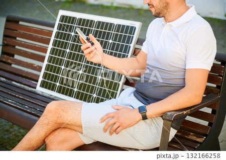 Cropped view of man using smartphone connected to photovoltaic solar panel. Integration of sustainable renewable energy into everyday life, showing practical use of solar power for charging devices. 132166258
