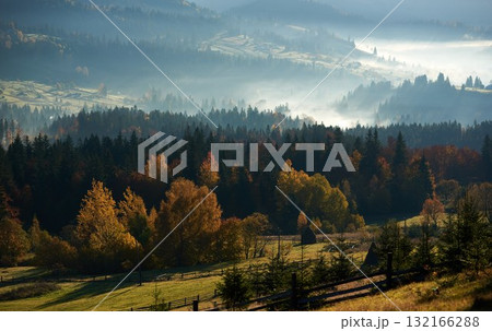 Autumn foliage in vibrant hues of gold, red, and orange blankets the forested hills, with mist rising gently in distance. Haystack in sunlit meadow, adding rustic charm to serene landscape. 132166288