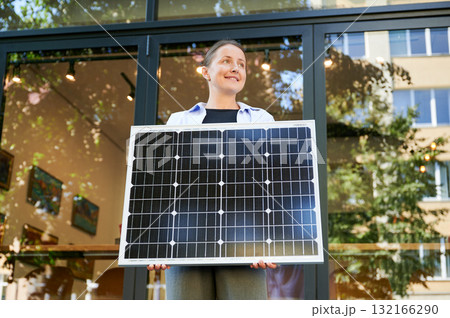 Woman holding solar panel for charging, stands outdoors, smiling in urban setting. Concept of green energy generation. 132166290