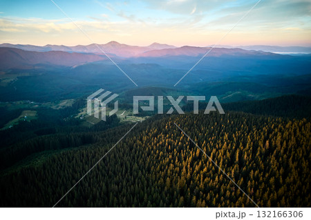 Aerial view of sunlit forested mountain with long shadows casting across vibrant green trees. Horizon stretches to distant mountains under clear blue sky, creating serene and expansive landscape. Aerial view of sunlit forested mountain with long shadows casting across vibrant green trees. Horizon stretches to distant mountains under clear blue sky, creating serene and expansive landscape. 132166306