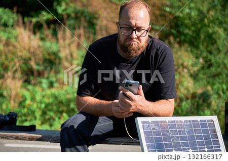 Bearded man using smartphone connected to photovoltaic solar panel. Integration of sustainable renewable energy into everyday life, demonstrating practical use of solar power for charging devices. Bearded man using smartphone connected to photovoltaic solar panel. Integration of sustainable renewable energy into everyday life, demonstrating practical use of solar power for charging devices. 132166317