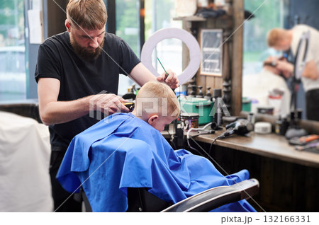 Hairdresser shaving boy's head. Competent barber using electric shaver at barbershop. Little client getting stylish men's haircut. 132166331