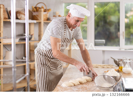 Elderly experienced male baker using knife to cut raw dough to make croissants Elderly experienced male baker using knife to cut raw dough to make croissants 132166666