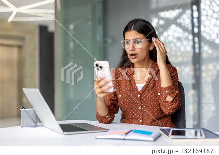 Young woman with glasses experiencing surprise and disbelief while reading upsetting information on her smartphone, sitting at an office desk with a laptop Young woman with glasses experiencing surprise and disbelief while reading upsetting information on her smartphone, sitting at an office desk with a laptop 132169187