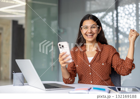 Young indian woman expressing excitement and happiness while looking at her smartphone, celebrating good news or achievement in a modern office, with a laptop and desk items Young indian woman expressing excitement and happiness while looking at her smartphone, celebrating good news or achievement in a modern office, with a laptop and desk items 132169188