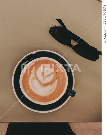 Coffee art and sunglasses on a table. A latte with beautiful coffee art sits on a saucer next to a pair of black sunglasses on a table, top-down view. Coffee art and sunglasses on a table. A latte with beautiful coffee art sits on a saucer next to a pair of black sunglasses on a table, top-down view. 132170678