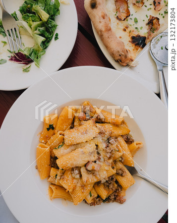 Rigatoni pasta with bolognese sauce, grated cheese and parsley served on white plate on restaurant table with pizza and green salad in background. Flatlay of pizza, rigatoni pasta and green salad Rigatoni pasta with bolognese sauce, grated cheese and parsley served on white plate on restaurant table with pizza and green salad in background. Flatlay of pizza, rigatoni pasta and green salad 132173804