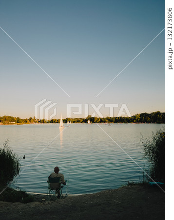 Man sitting on folding chair by lake, fishing and enjoying view of sailboats gliding across water during stunning sunset, embracing tranquility and relaxation. Fisherman on lakeside folding chair 132173806