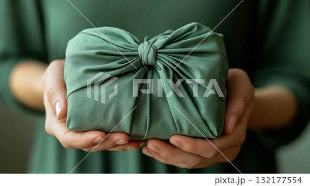 Hands holding green fabric-wrapped gift in eco-friendly style on soft background. Christmas decor and craftwork, eco friendly, sustainable concept. 132177554